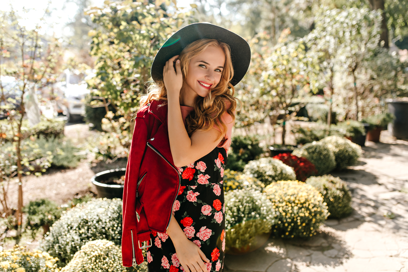 beautiful-woman-in-wide-brimmed-hat-coquettishly-touches-hair-woman-in-red-jacket-and-dress-with-roses-is-smiling-in-garden